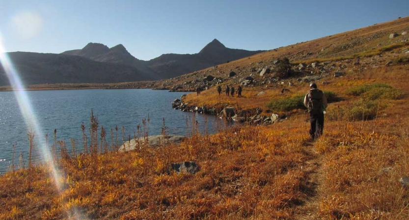 A group of people walk on a trail alongside a blue alpine lake. There are mountains in the background. 